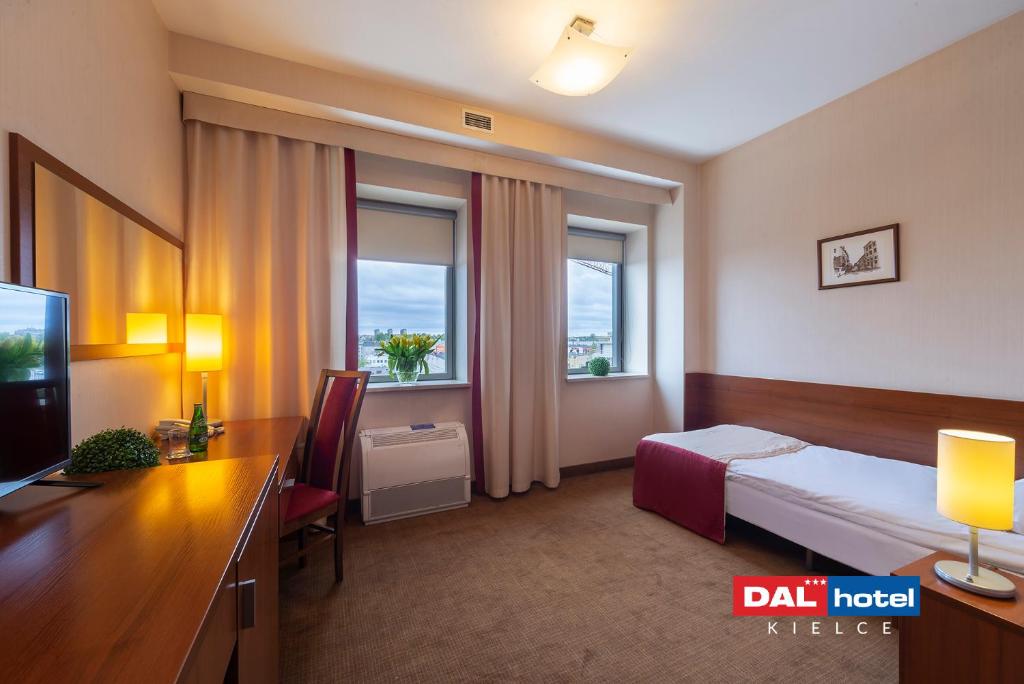 Compact single room with wooden desk, bedside table, lamp, burgundy bed skirt and two windows.