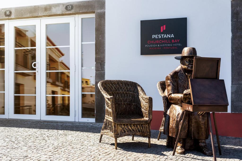 Una statua di un uomo seduto su una sedia di fronte a un edificio di Pestana Churchill Bay a Câmara de Lobos