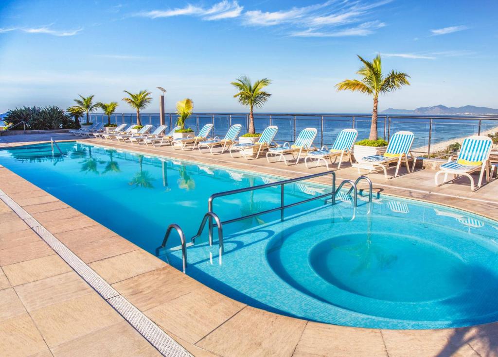 einen Pool mit Stühlen und das Meer im Hintergrund in der Unterkunft Windsor Barra Hotel in Rio de Janeiro