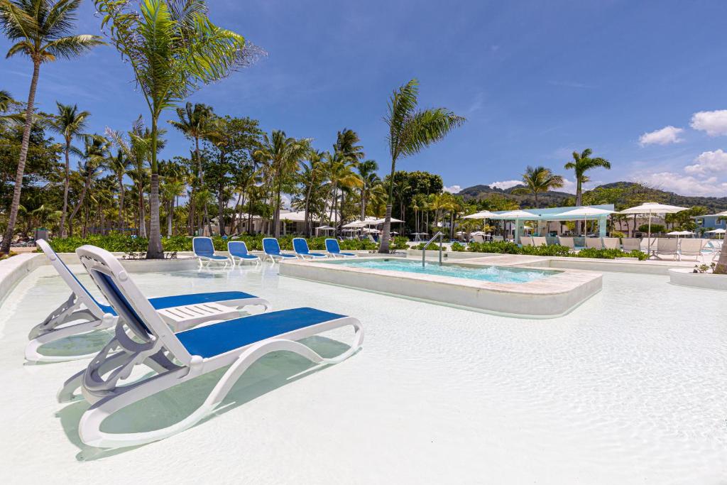 Outdoor pool deck with loungers, palm trees, umbrellas, and mountainous landscape backdrop.
