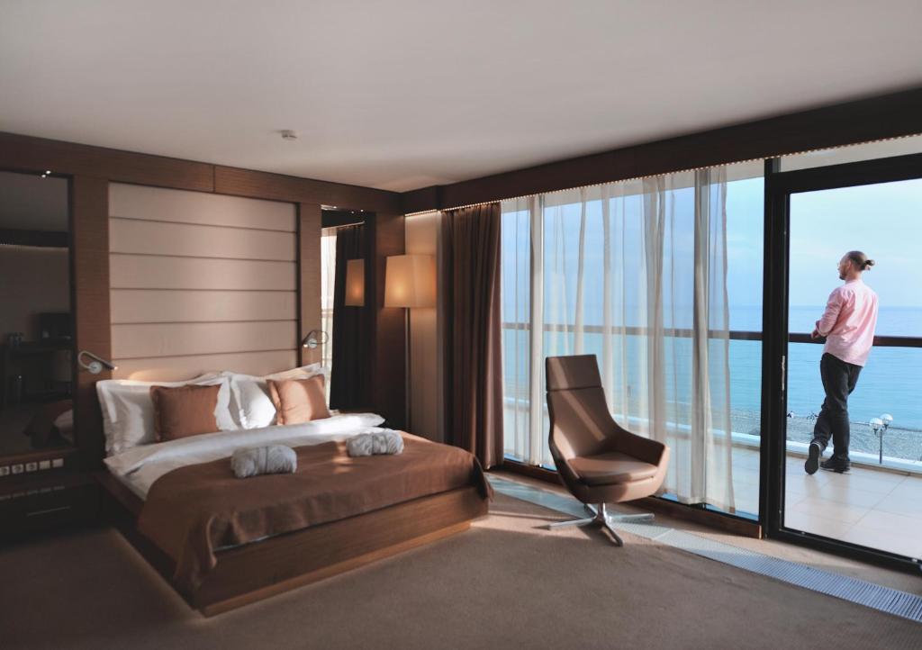 a man standing in a bedroom with a bed and a chair at Arfa Park-Hotel in Adler