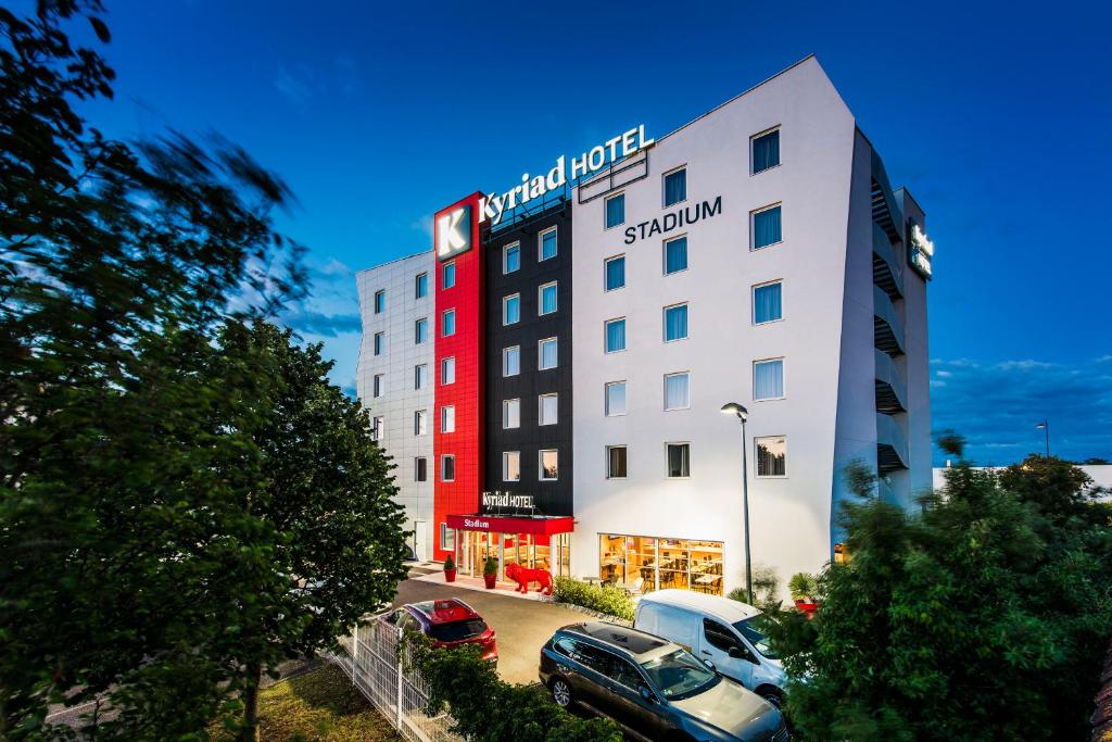 un bâtiment d'hôtel avec des voitures garées dans un parking dans l'établissement Kyriad Lyon Est Stadium Eurexpo Meyzieu, à Meyzieu