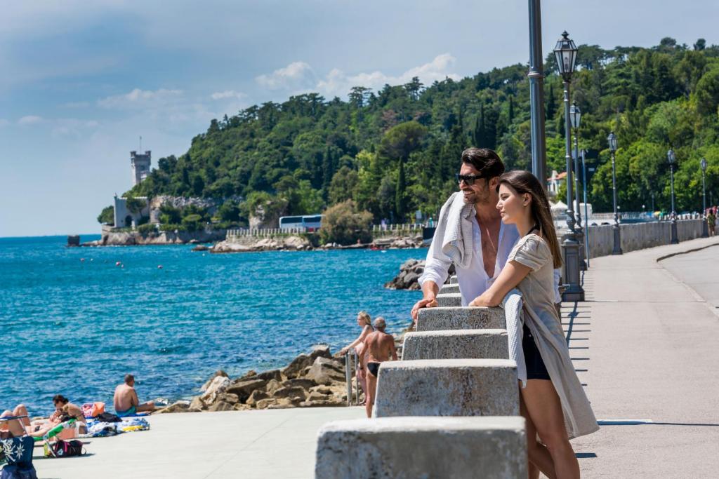 a man and a woman standing on a sidewalk near the water at Hotel Miramare - Adults Only in Trieste