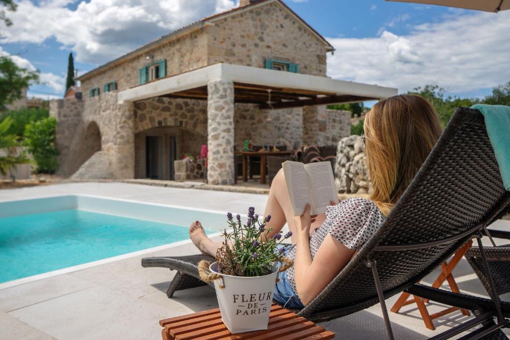 a woman sitting in a chair reading a book by a pool at Vila na Kamenu in Dobrinj