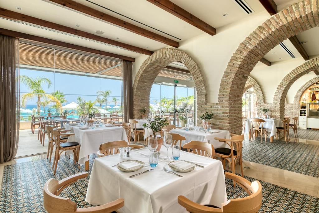 un restaurant avec des tables et des chaises blanches et de grandes fenêtres dans l'établissement Royal Apollonia by Louis Hotels, à Limassol