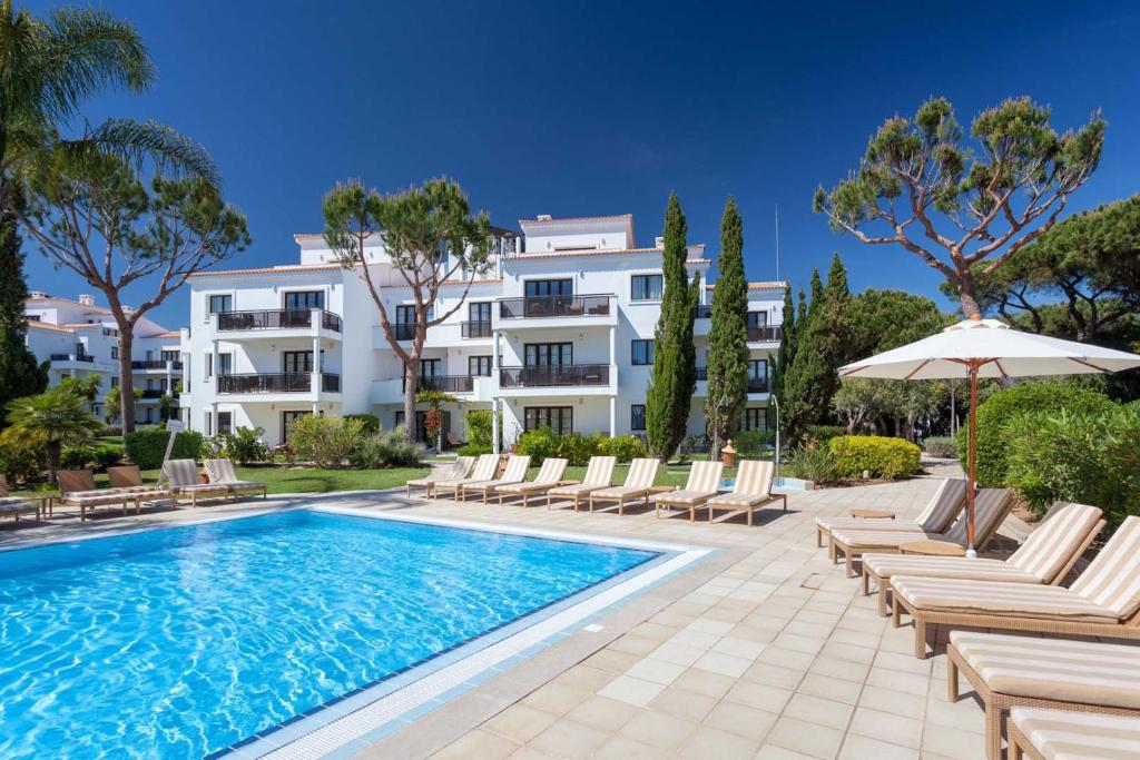 a swimming pool with chairs and an umbrella and a building at Pine Cliffs Village & Golf Suites in Albufeira