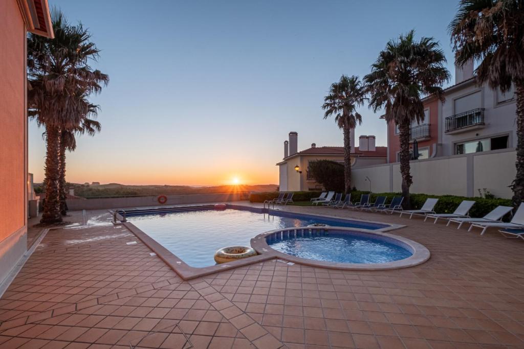 a pool in a courtyard with palm trees and a sunset at Acolhedora Vivenda Geminada Praia D'el Rey in Casal da Lagoa Seca