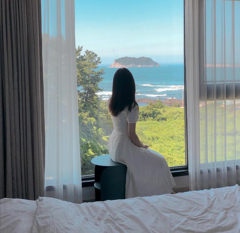 Una mujer con un vestido blanco mirando por una ventana al océano en Hotel Seogwipean en Seogwipo