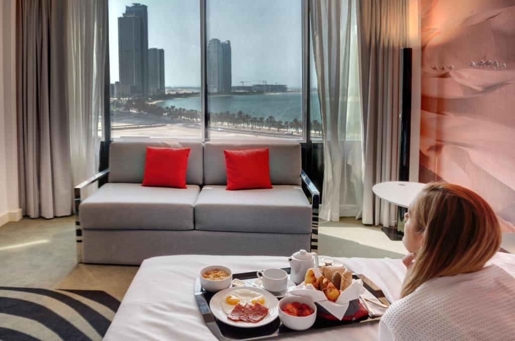 a woman sitting in a hotel room with a tray of food at Novotel Sharjah Expo Centre in Sharjah