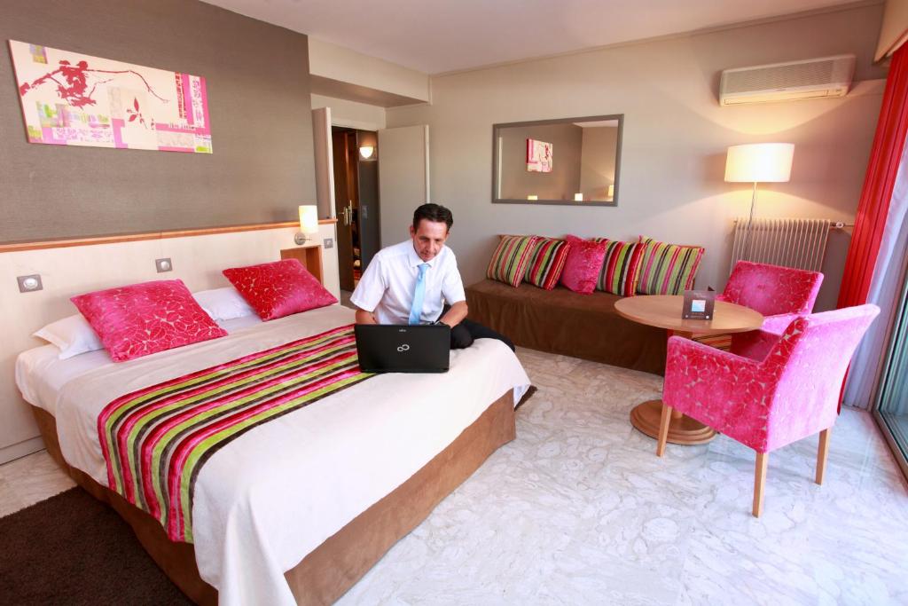 a man sitting on a bed in a hotel room with a laptop at The Originals Boutique, Hôtel Les Strélitzias in Juan-les-Pins