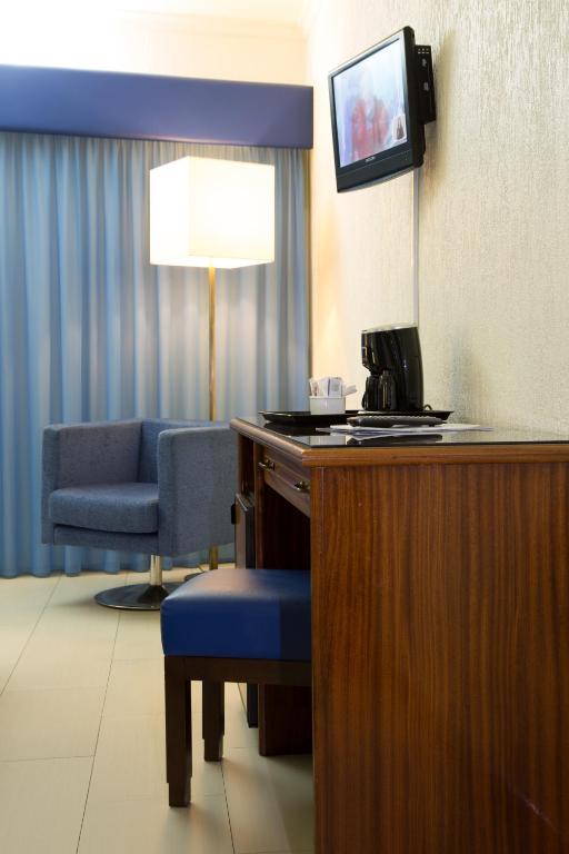 A compact in-room desk setup with a wall-mounted television, coffee machine and upholstered chair.