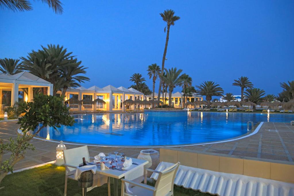 einen Pool in der Nacht mit einem Tisch, Stühlen und Palmen in der Unterkunft Djerba Golf Resort & Spa in Midoun