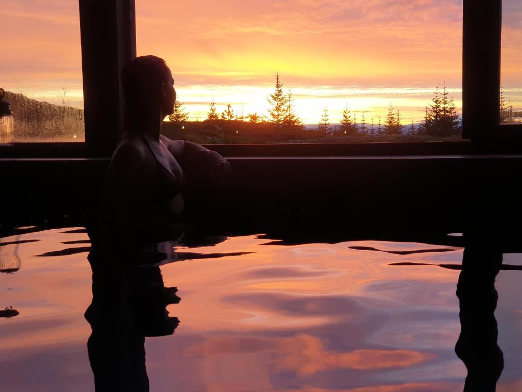 uma mulher sentada numa banheira a olhar pela janela ao pôr-do-sol em 360 Hotel & Thermal Baths em Selfoss