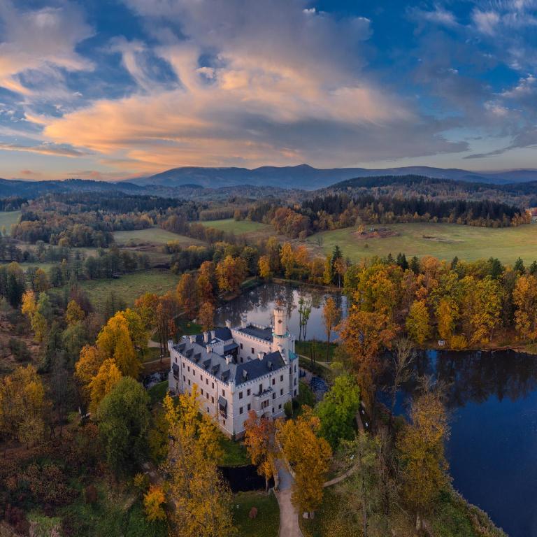 Aerial view of white lakeside castle surrounded by autumn trees, rolling hills and dramatic sunset sky.