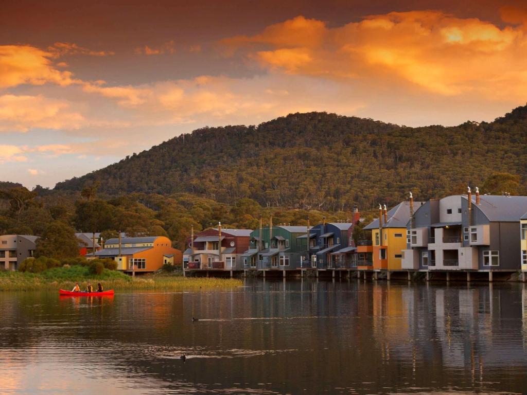 a small town on the water with houses and a boat at Novotel Lake Crackenback Resort in Crackenback