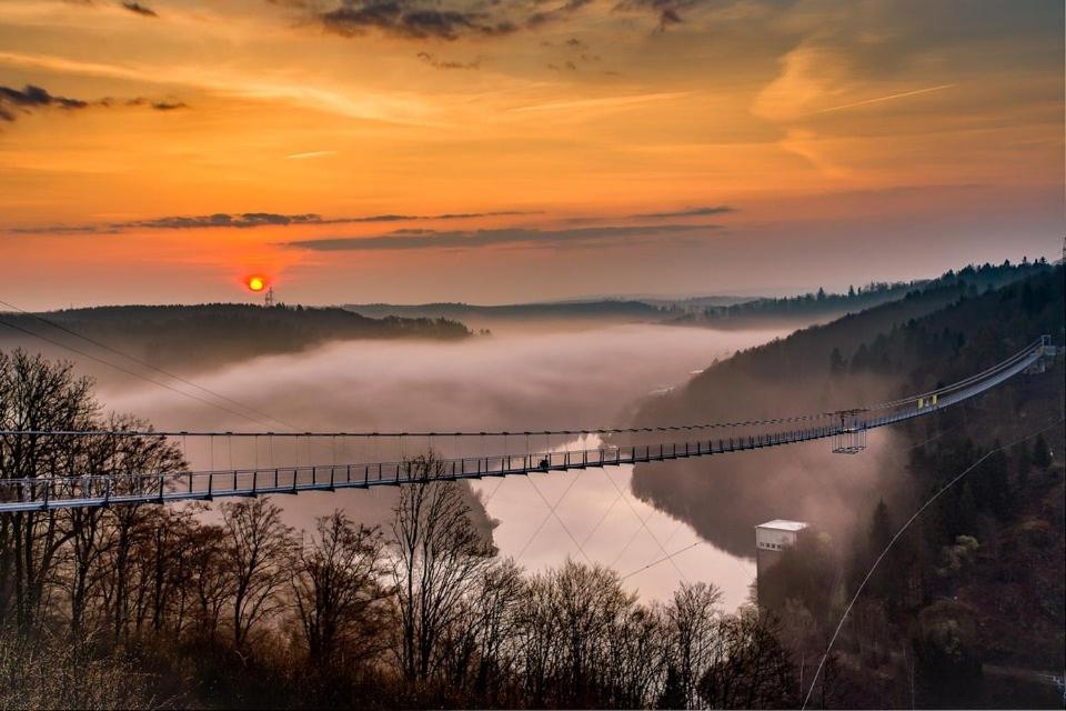 een hangbrug over een mistige rivier met de zon op de achtergrond bij Ferienwohnung Am Eichenberg in Blankenburg