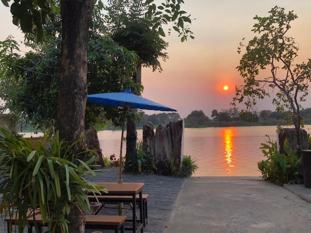 un coucher de soleil sur l'eau avec des tables et un parasol dans l'établissement Ruen Thai Ban Rim Nam, à Kamphaeng Phet