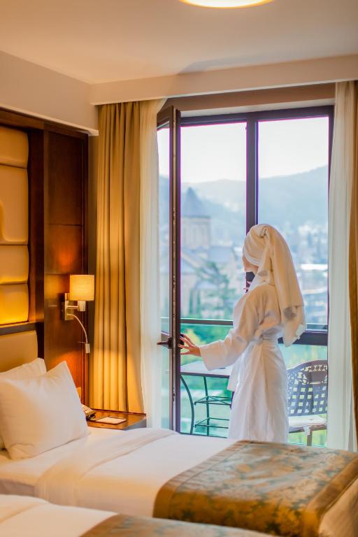 a woman in a white robe looking out the window of a hotel room at KMM Hotel in Tbilisi City