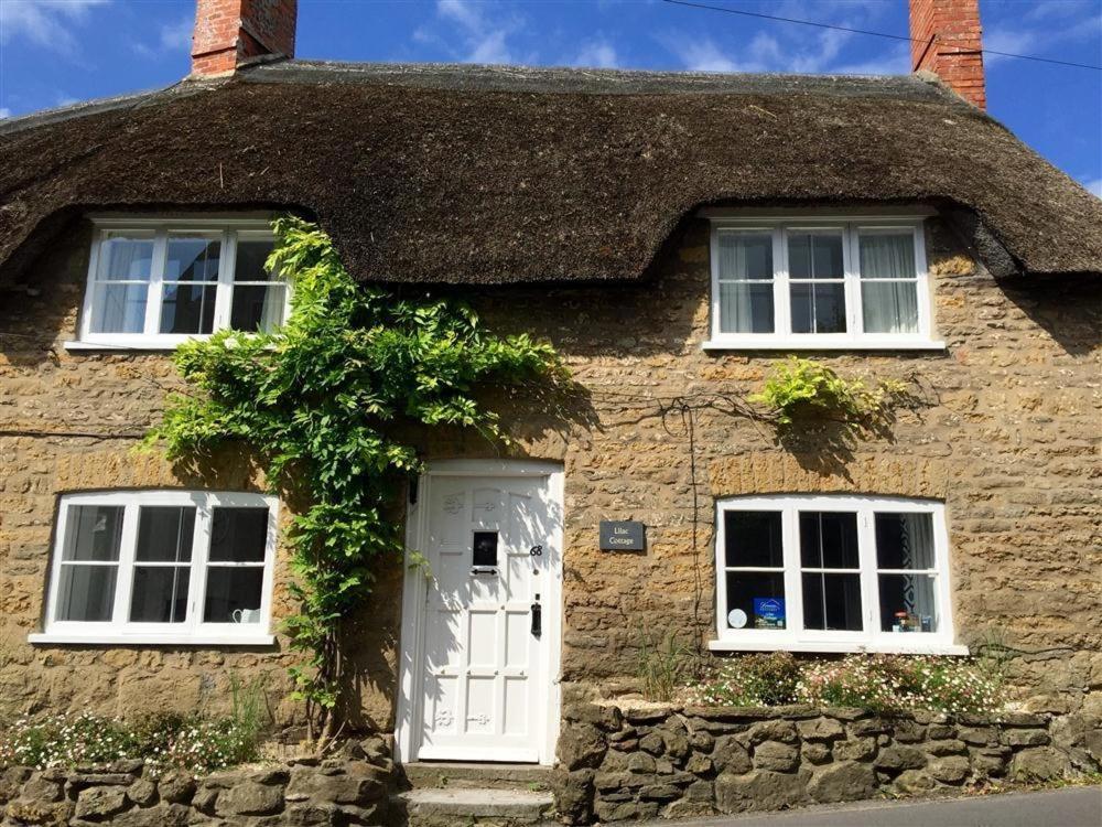 a thatched cottage with a white door and two windows at Lilac Cottage in Burton Bradstock