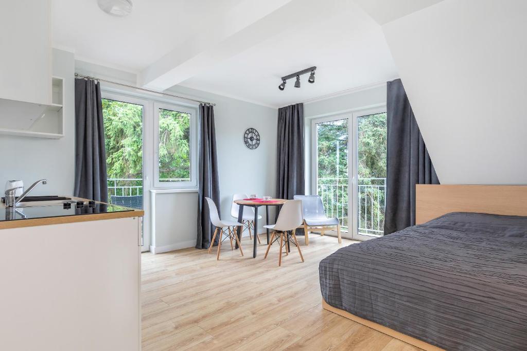 Studio bedroom featuring bed with dark bedding, small dining table, kitchenette and large windows overlooking trees.