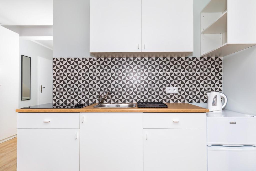Compact white kitchenette with wooden countertop, sink, stovetop and black-and-white geometric tile backsplash, kettle visible.