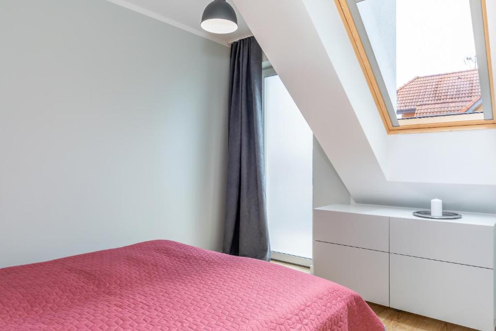 Minimal bedroom corner with red bedspread, slanted skylight, gray curtains, white dresser topped with candle.