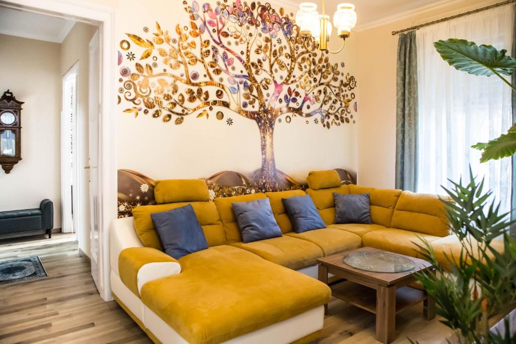 a living room with a yellow couch with a tree on the wall at Villa Apartman in Makó