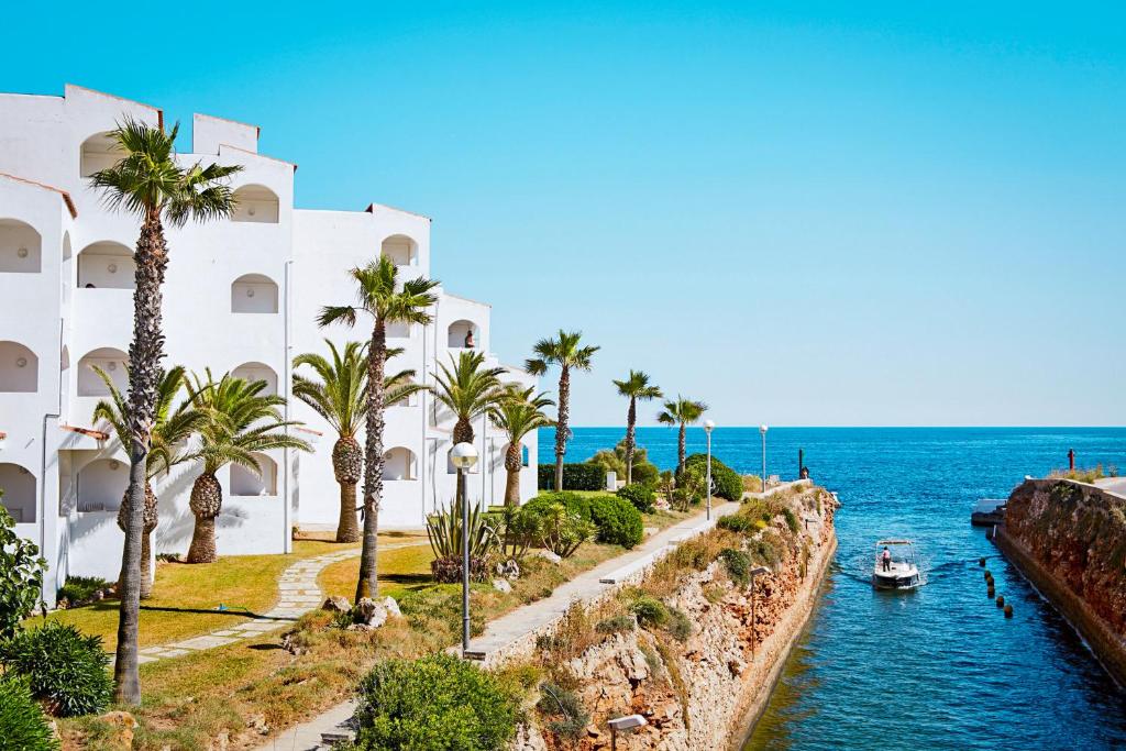 a building with palm trees and a boat in the water at Globales Apartamentos Marina in Cala en Bosc