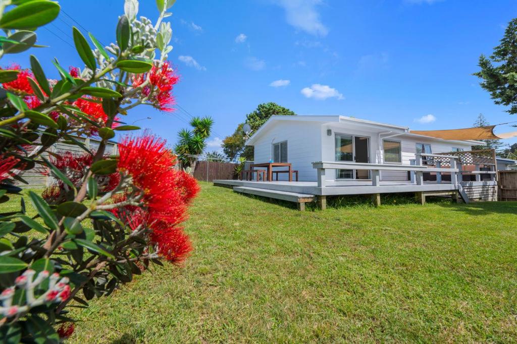 a small white house in a yard with red flowers at Pohutukawa Breeze - Papamoa Beach Holiday Home in Papamoa