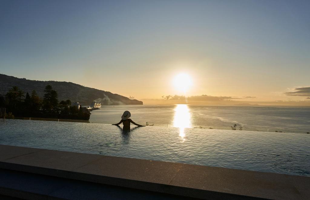 a person sitting in the water in a pool with the sunset at Les Suites at The Cliff Bay - PortoBay in Funchal
