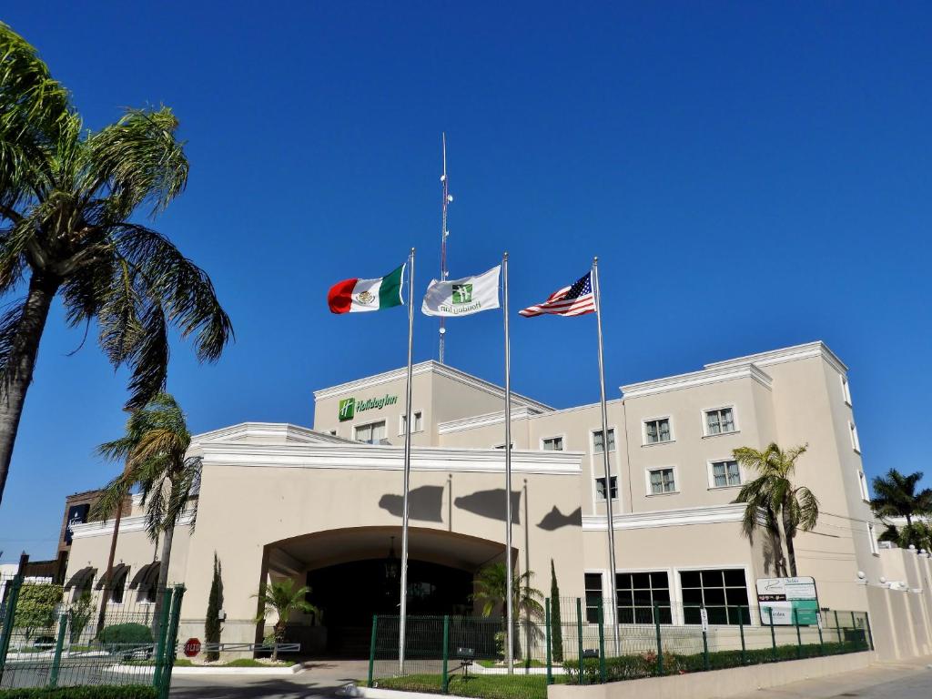 three flags flying in front of a building with palm trees at Holiday Inn Reynosa Zona Dorada by IHG in Reynosa