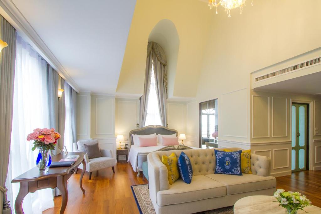 A bright high-ceilinged room with a bed, sofa, writing table and tall curtained windows.