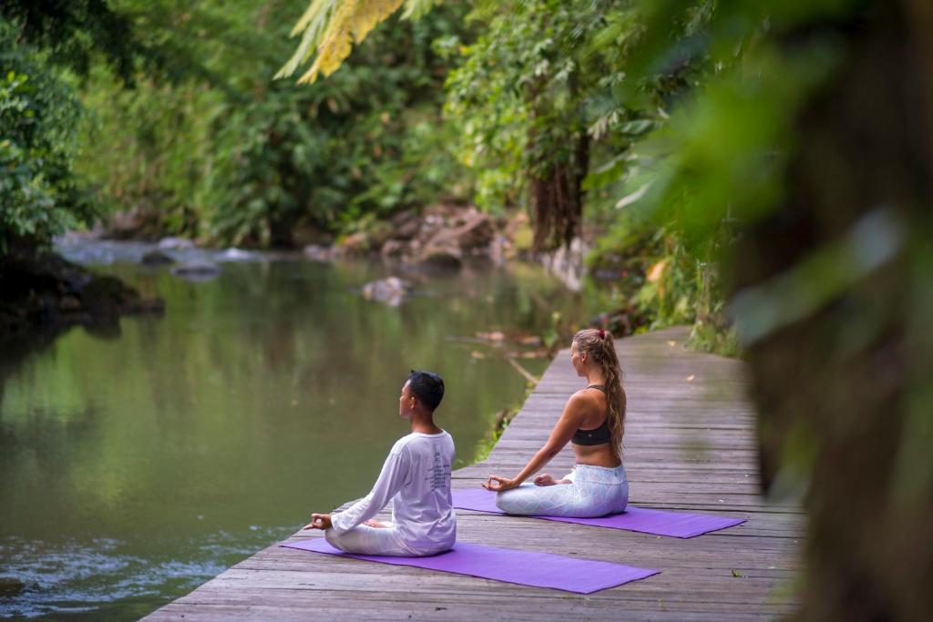 a man and woman sitting on a dock doing yoga by a river at Adiwana Svarga Loka - A Retreat Resort in Ubud