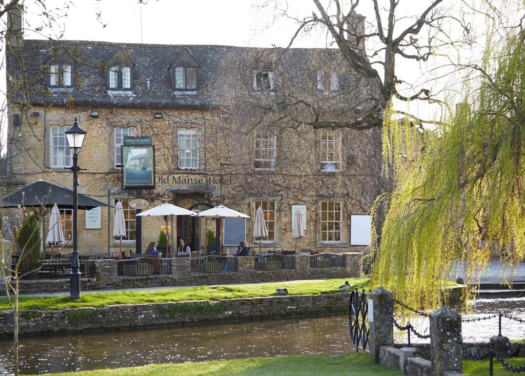 a large stone building with tables and umbrellas next to a river at Old Manse Hotel by Greene King Inns in Bourton on the Water