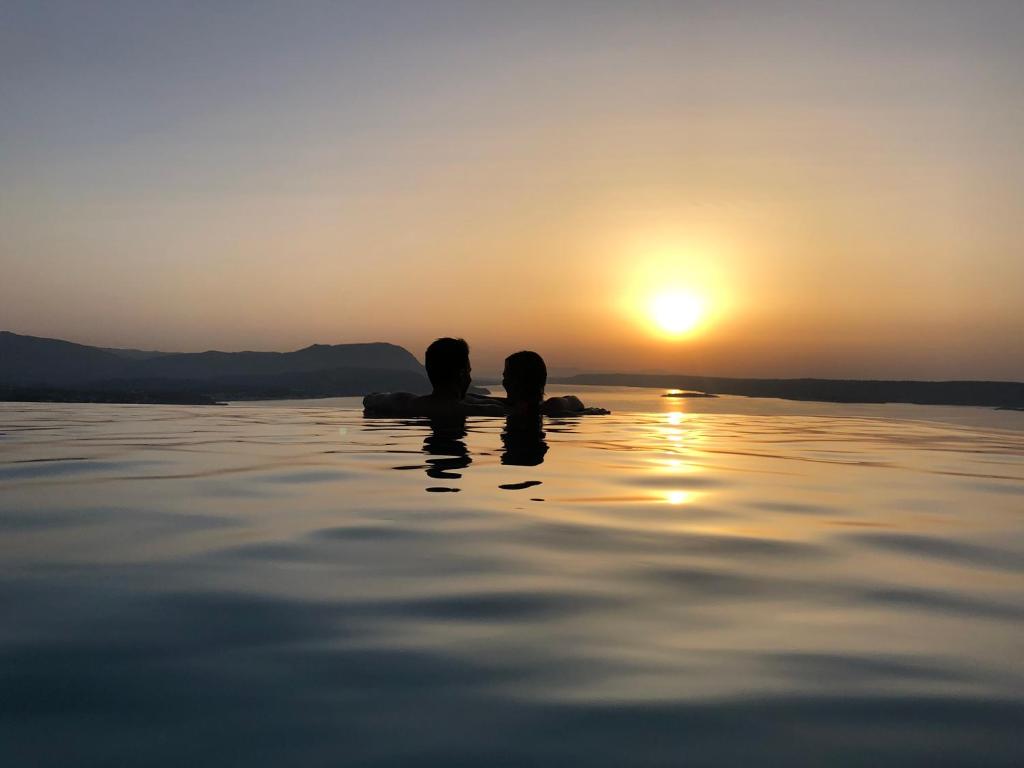 Due persone sedute in acqua a guardare il tramonto di PhantΩm Villa a Kókkinon Khoríon