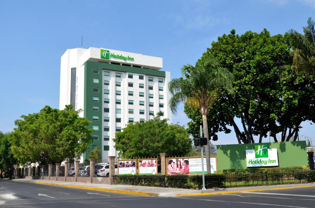 Holiday Inn Guadalajara Expo Plaza del Sol, an IHG Hotel في غواذالاخارا: مبنى اخضر وابيض وبه اشجار وشارع