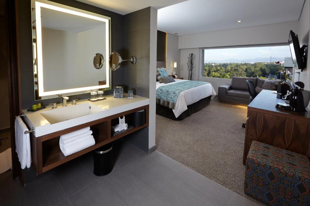 A hotel room viewed from its open vanity area, with a bed, sofa and broad green outlook.
