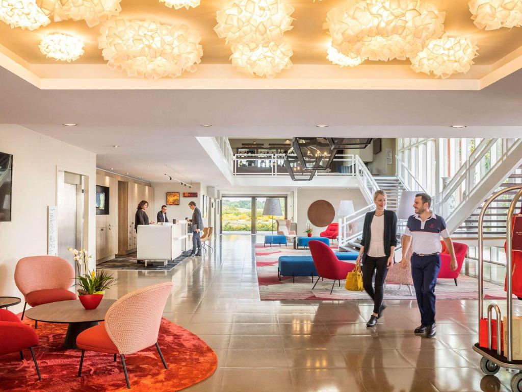 2 personnes traversant un hall avec des chaises rouges dans l'établissement Golf du Médoc Resort Bordeaux - MGallery, à Le Pian-Médoc