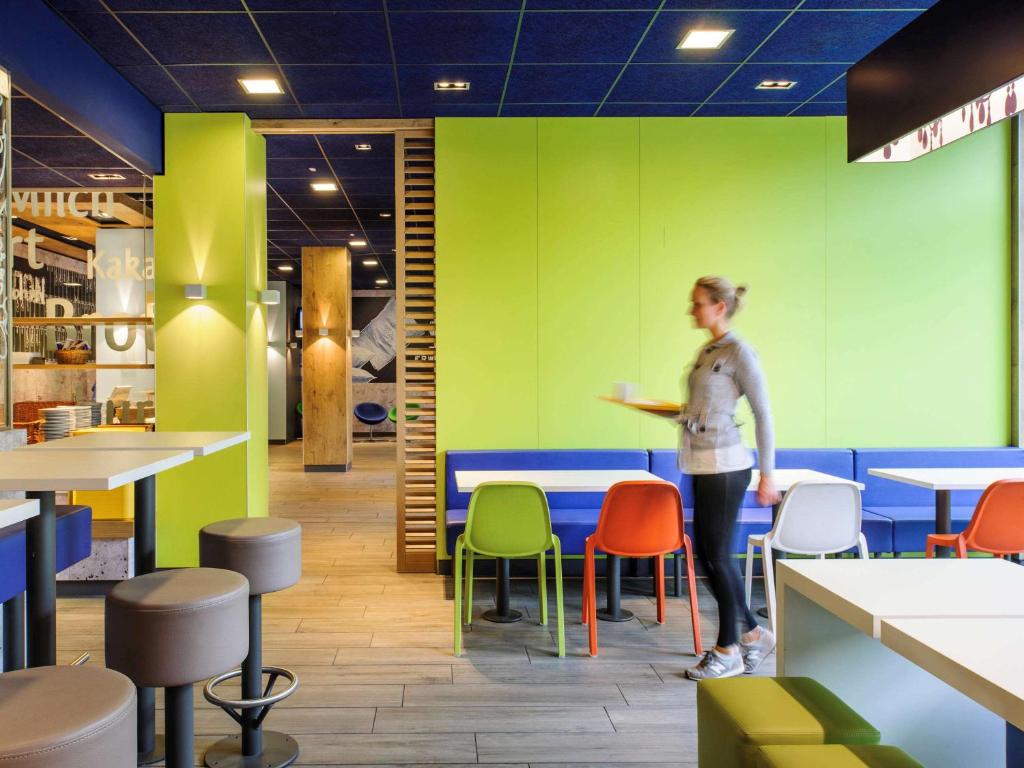 Hotel ibis Budget Laval في Bonchamp-lès-Laval: امرأة تقف في مطعم بألوان زاهية مع الطاولات والكراسي
