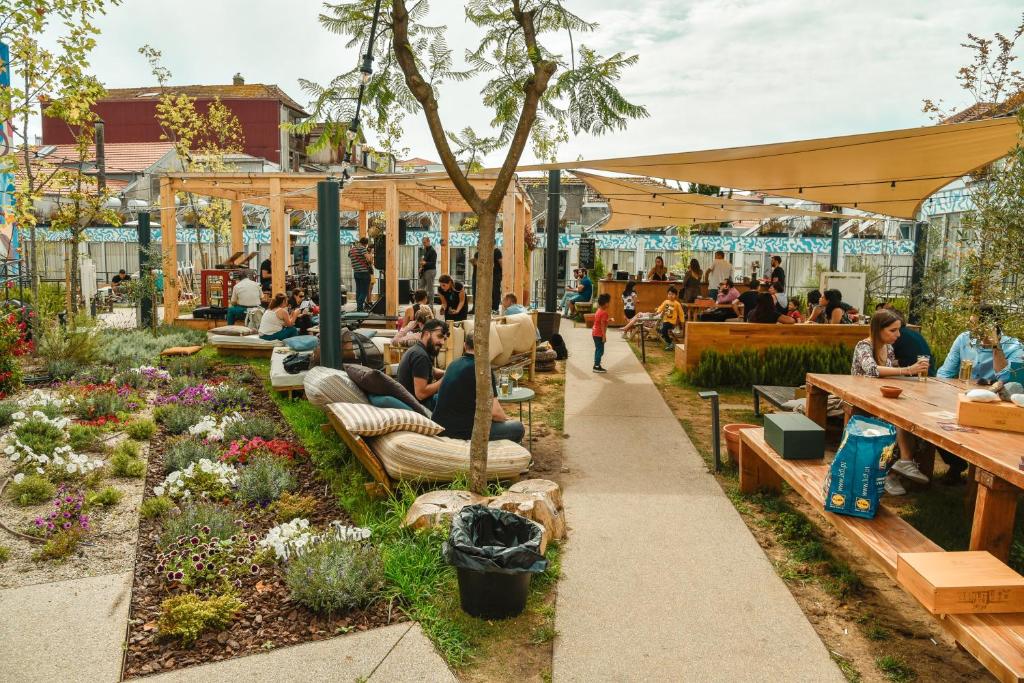 Outdoor garden lounge at Selina Porto featuring wooden benches, picnic tables, and hammock seating surrounded by raised garden beds and young trees, with guests relaxing under canvas shade sails.