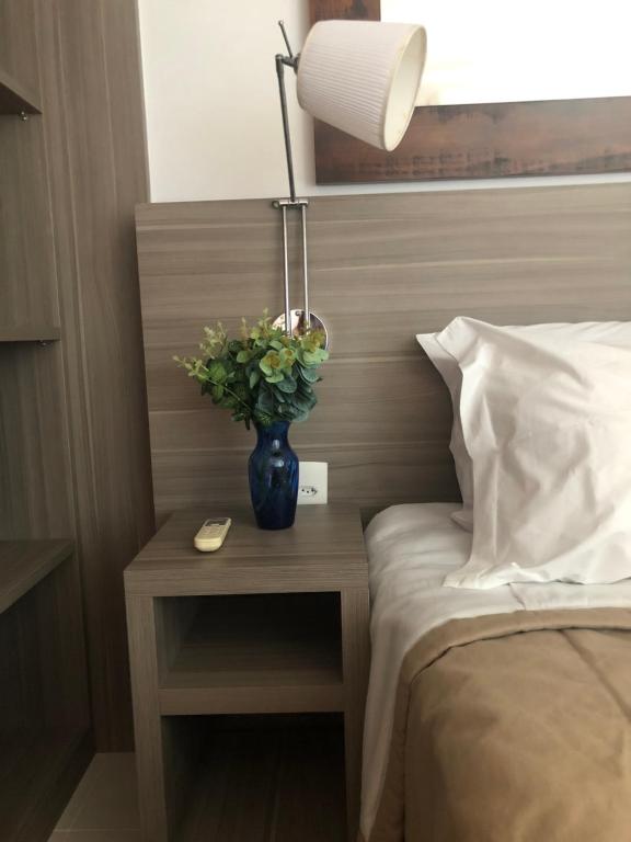 einen Tisch mit einer Lampe und eine Vase Blumen auf einem Bett in der Unterkunft Flat 507 próximo ao Projac Jacarepaguá in Rio de Janeiro