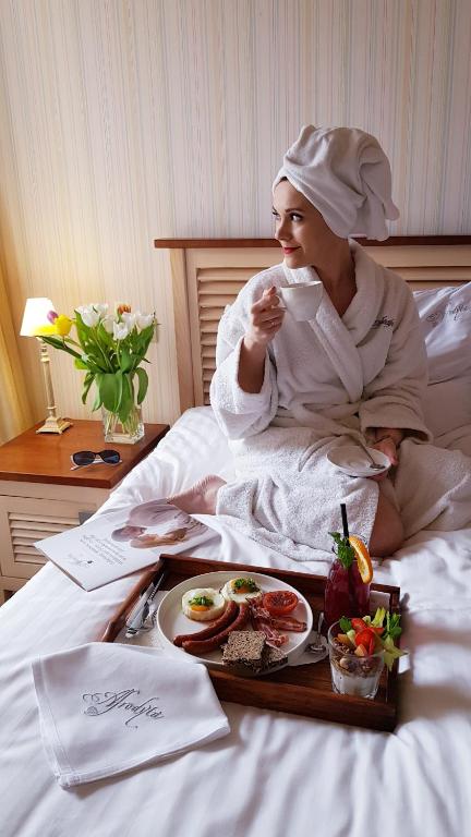 Person in bathrobe enjoying breakfast in bed, with flowers and a lamp on bedside table.