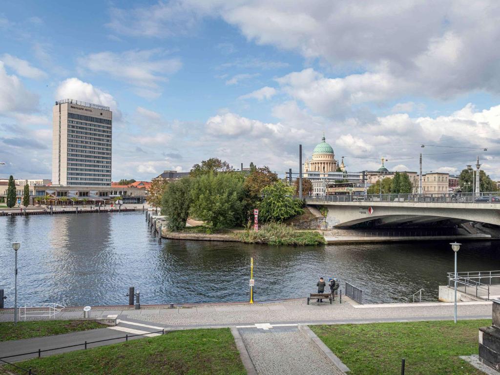 una persona sentada en un banco junto a un río con un puente en Mercure Hotel Potsdam City, en Potsdam