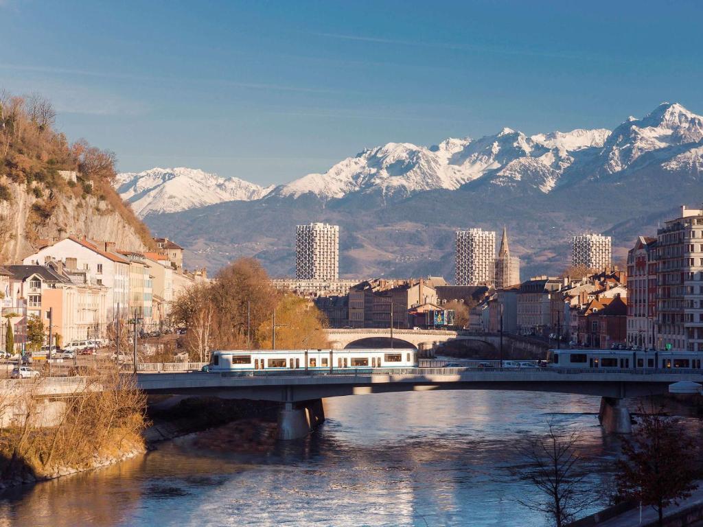 a train on a bridge over a river with snow covered mountains at ibis Grenoble Gare in Grenoble