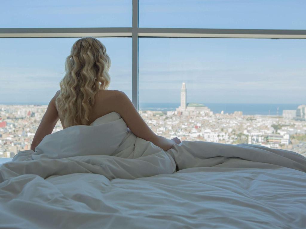 una mujer sentada en una cama mirando por la ventana en Sofitel Casablanca Tour Blanche en Casablanca