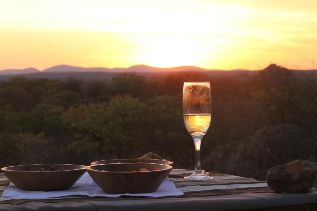 een glas wijn en twee kommen op een tafel met zonsondergang bij Kaoko Bush Lodge in Kamanjab