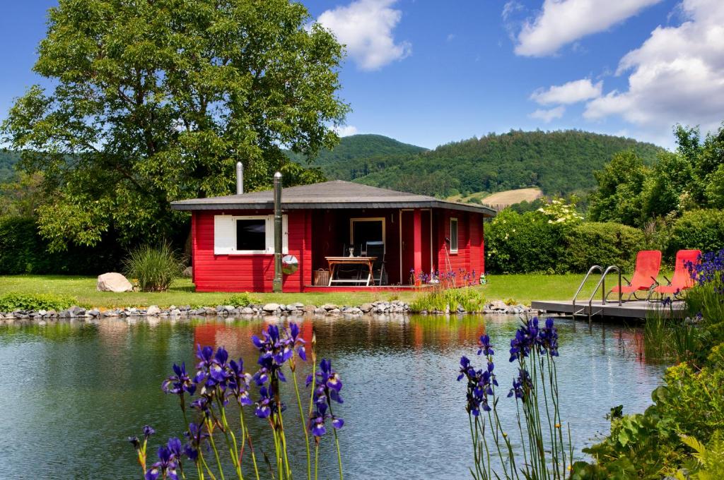 a red cabin on a lake with purple flowers at Romantic Wooden Lodge,Sauna,Schwimmteich,alleinstehend,absolut ruhig in Hessisch Oldendorf