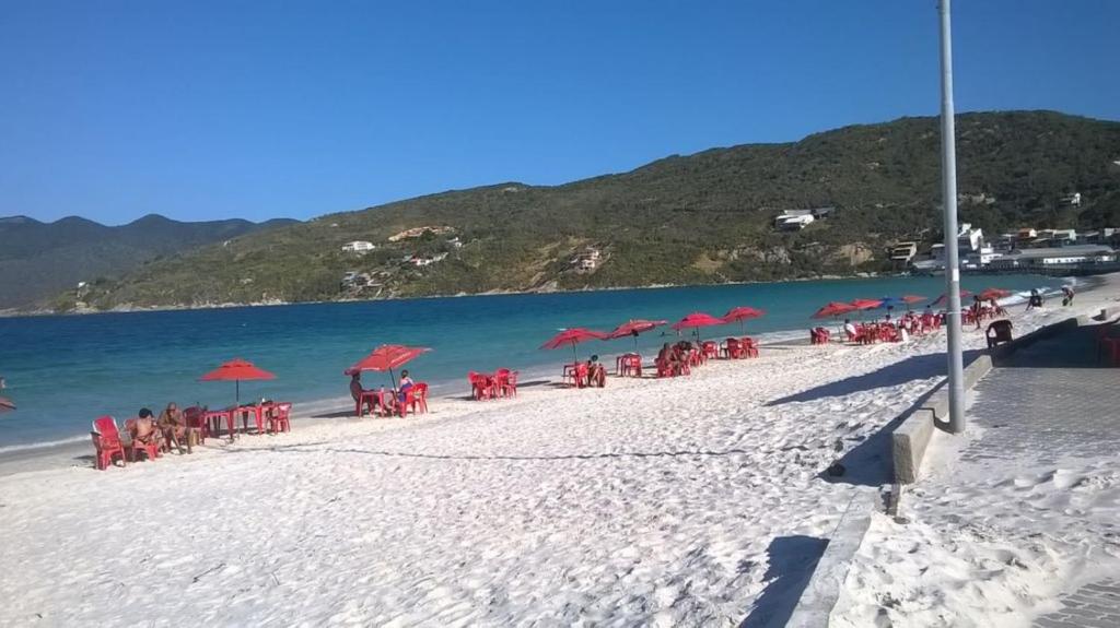 una playa con sillas, sombrillas rojas y agua en Apartamento Elizabetta 3, pé na areia, en Arraial do Cabo