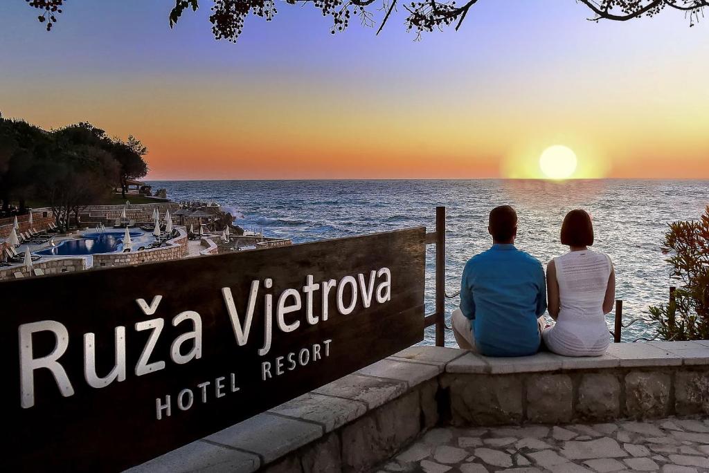 two people sitting on a sign looking at the ocean at Ruza Vjetrova - Wind Rose Hotel Resort in Dobra Voda