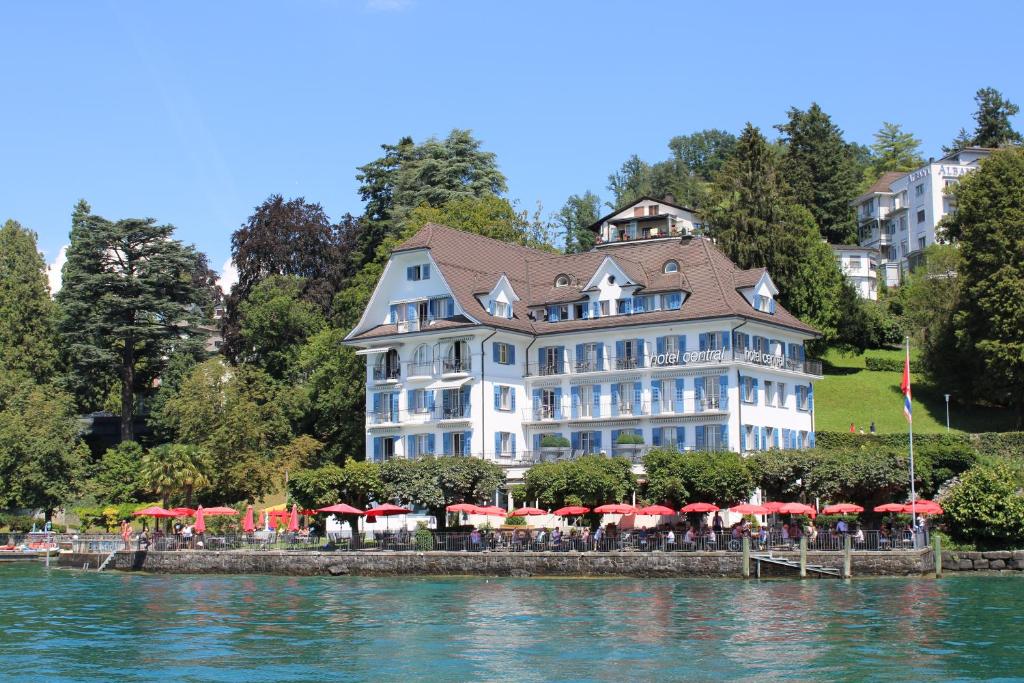 un grand bâtiment blanc avec des parasols rouges en face de l'eau dans l'établissement Hotel Central Am See - Beau Rivage Collection, à Weggis